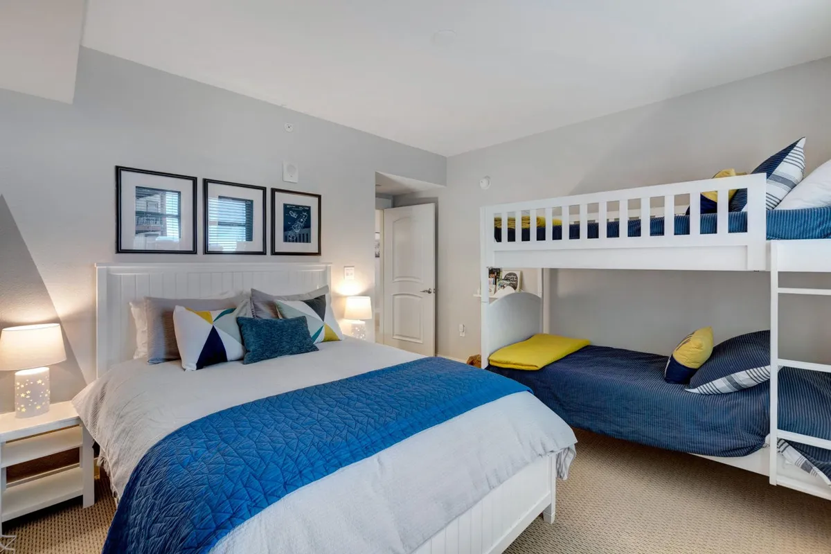 A cozy and family-friendly bedroom with a large bed dressed in blue and gray bedding alongside a twin-over-twin bunk bed with playful yellow accents. Framed artwork and soft lighting create a welcoming, multi-use space for children or additional guests.