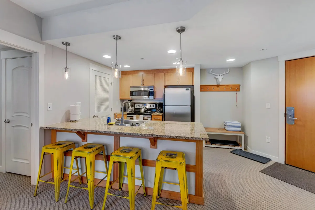 A cozy kitchen scene with a granite countertop, cheerful yellow bar stools, and stainless steel appliances. The wooden accent wall adds warmth and a homely touch, while the layout encourages gathering and social interaction during cooking or casual meals.