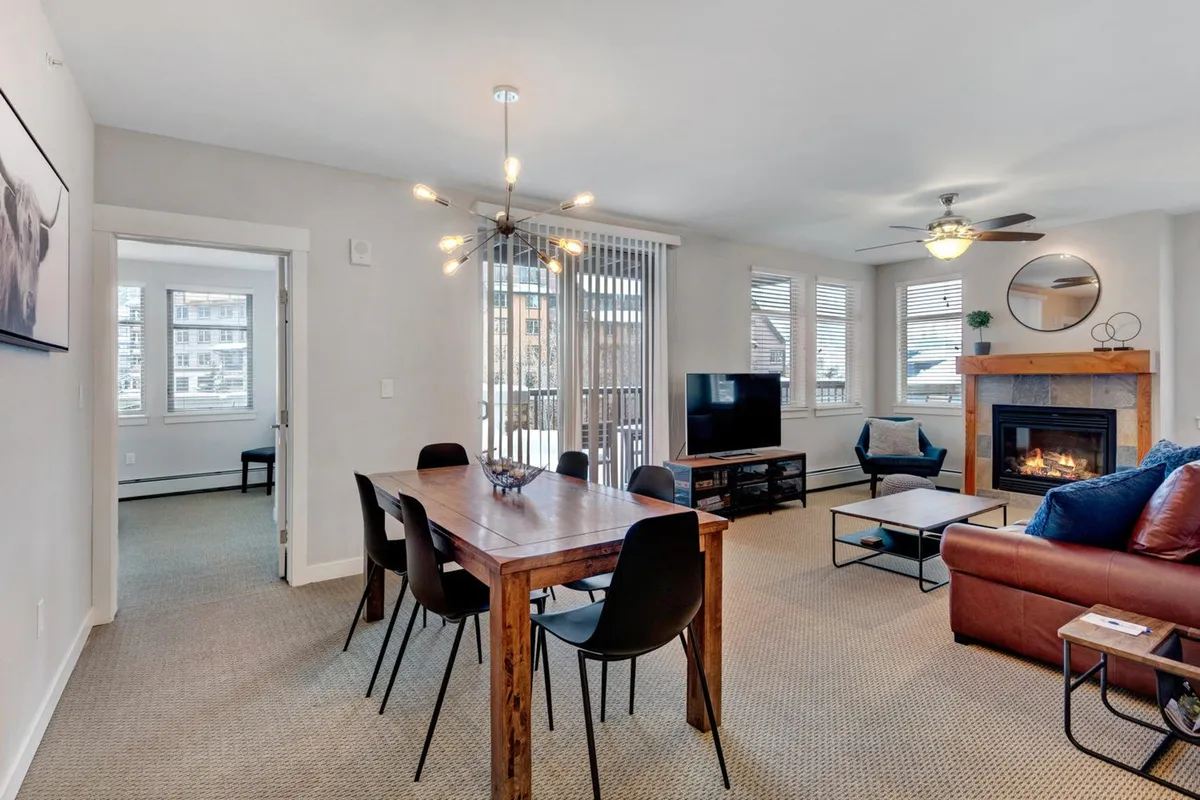 A spacious open-plan living and dining area highlighting a comfortable seating area, a prominent fireplace, a modern dining table, and a wall-mounted TV. Large windows fill the room with natural light and offer glimpses of the mountain landscape outside.