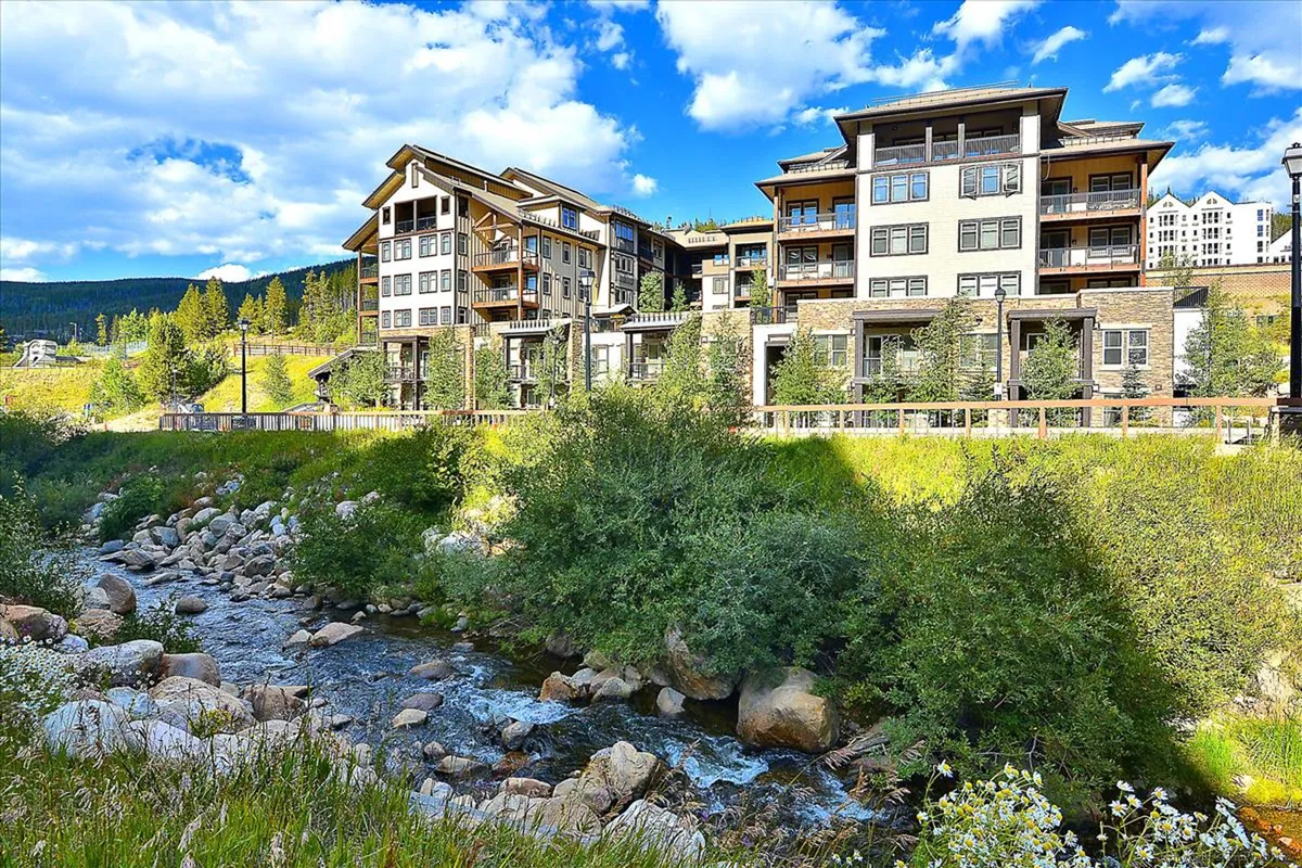 A modern resort building stands against a backdrop of dramatic mountains, bordered by lush greenery and a sparkling stream. Outdoor seating areas invite guests to relax and take in the sunset views, with nature trails and wildflowers visible nearby.