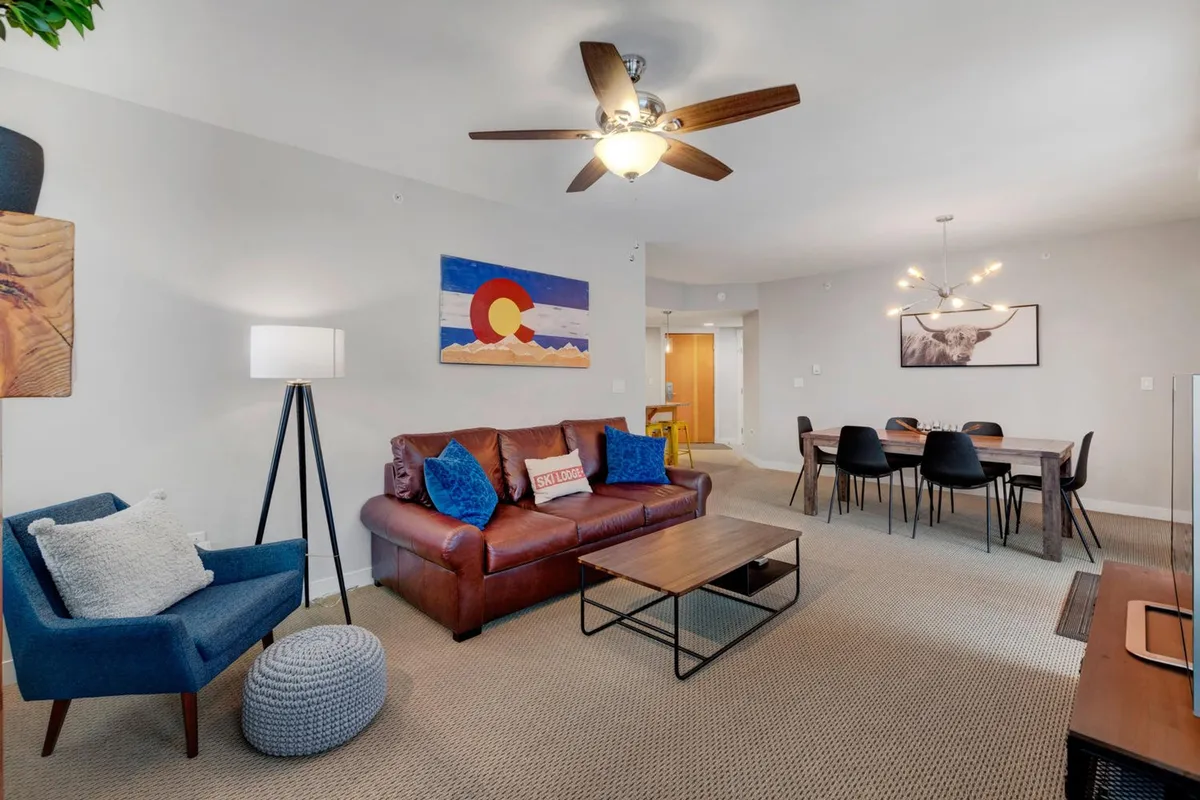 A view of the living room showing a comfortable sofa, a dining table set for gatherings, stylish artwork, and soft lighting from a modern lamp. The space is designed for leisure and socializing, and the sofa can double as a sleeper for extra guests.