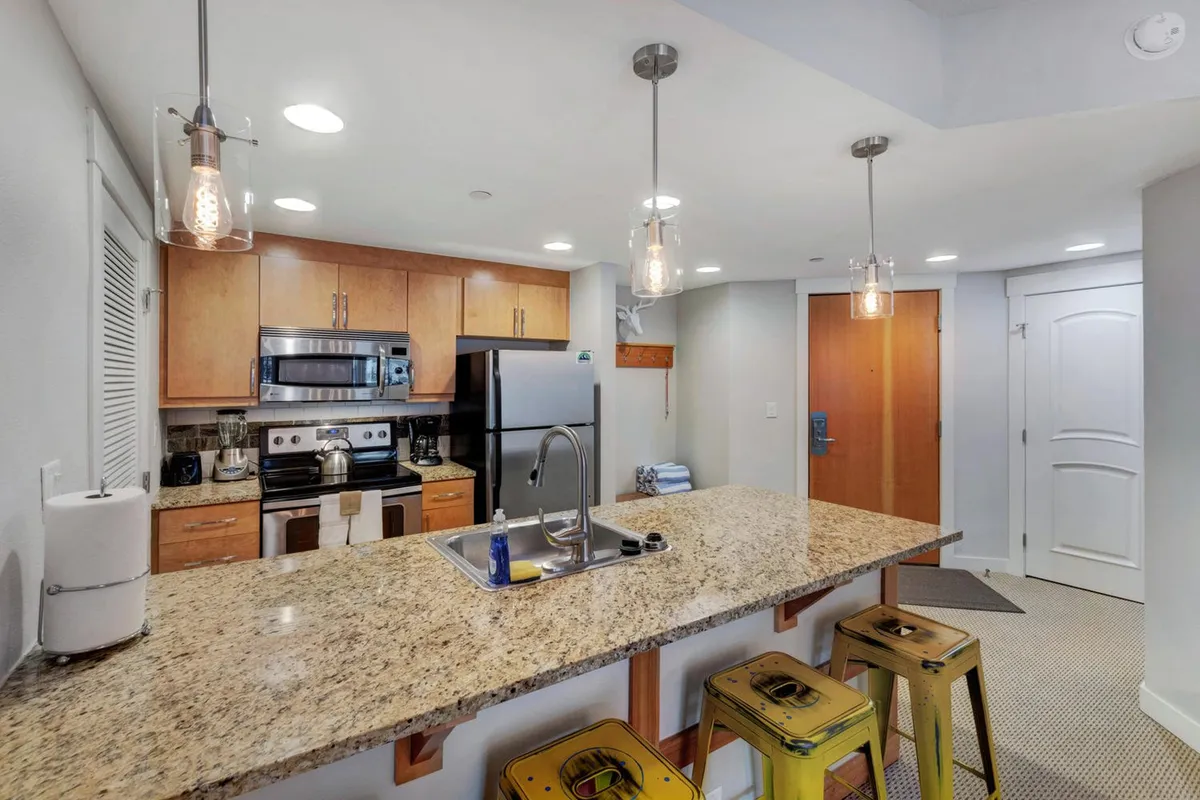A spacious, modern kitchen featuring gleaming granite countertops, stainless steel appliances, and vibrant pendant lights hanging above colorful bar stools. The open-concept layout invites both cooking and casual dining, while stylish fixtures and abundant prep space create an atmosphere suited for both culinary adventures and quick breakfasts.