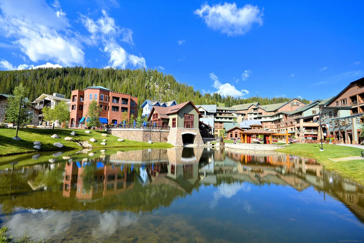 A picturesque exterior shot of colorful buildings reflected in a serene pond, with a bridge connecting pathways surrounded by trees. People stroll, picnic, and enjoy outdoor dining, creating a cheerful and relaxed village atmosphere that’s perfect for unwinding after a day of adventures.