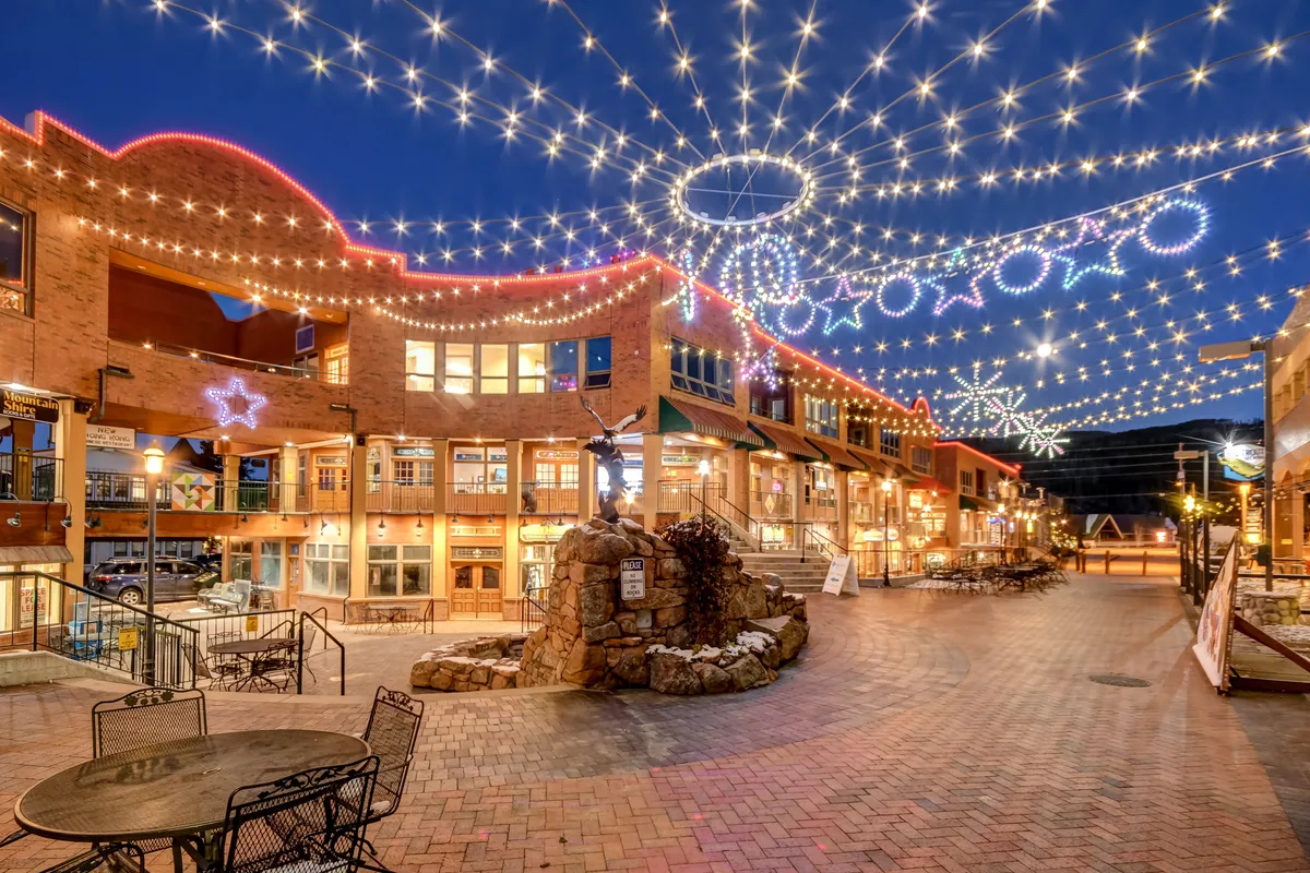 The photo captures a lively downtown street scene, illuminated by decorative lights. Outdoor seating, a stone fountain, and bustling storefronts suggest a vibrant atmosphere perfect for dining, shopping, and socializing throughout the year. Community gatherings and celebrations are hinted at by the festive mood.