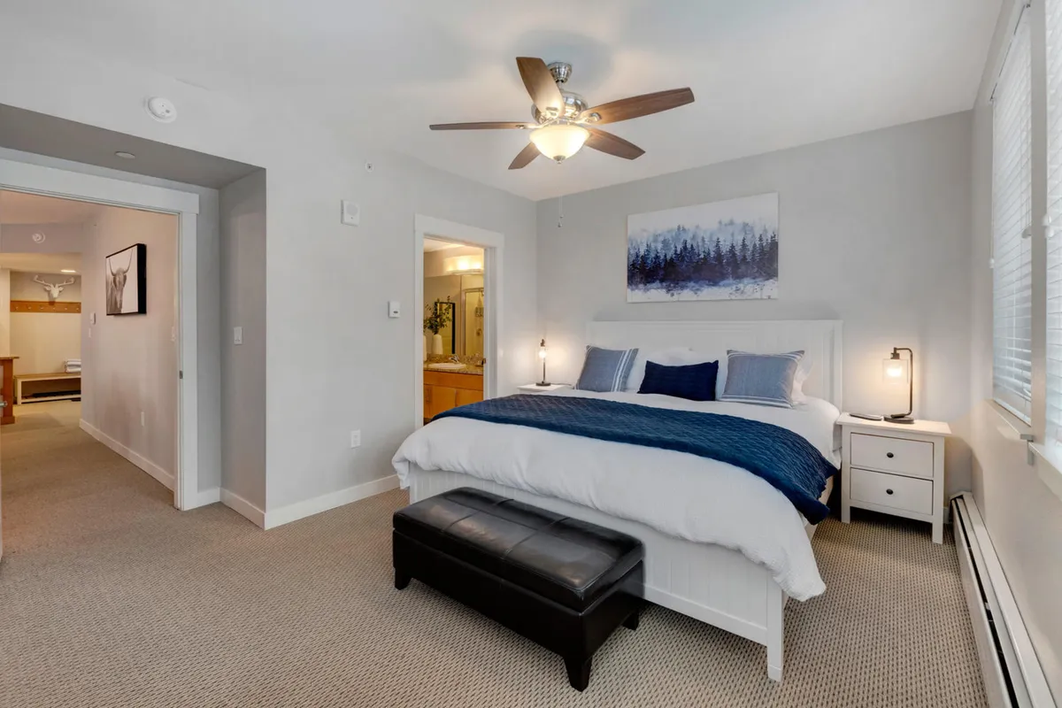 A tranquil bedroom featuring a large bed with plush bedding, a calming painting above the headboard, a nightstand with a lamp, and a small bench at the foot of the bed. The soft lighting and uncluttered decor evoke a relaxing retreat ideal for restful sleep.